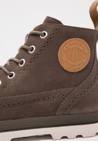 Botte montante en daim marron avec œillets argentés, motifs perforés, un patch en cuir beige marqué "PALLADIUM", et une semelle en caoutchouc texturée.