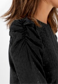 Femme portant un haut noir à manches longues avec des détails froncés aux épaules bouffantes et une texture florale subtile, avec des cheveux bruns visibles.