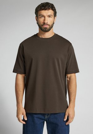 Mann mit lockigem Haar und Bart, der ein schlichtes dunkelbraunes kurzärmeliges T-Shirt und blaue Jeans vor neutralem Hintergrund trägt.