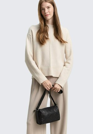 Schwarze gesteppte Handtasche mit einem Tragegurt, in einer Hand gehalten. Cremefarbener Strickpullover und hellbeige Hose, getragen von einem Modell vor einem grauen Hintergrund.