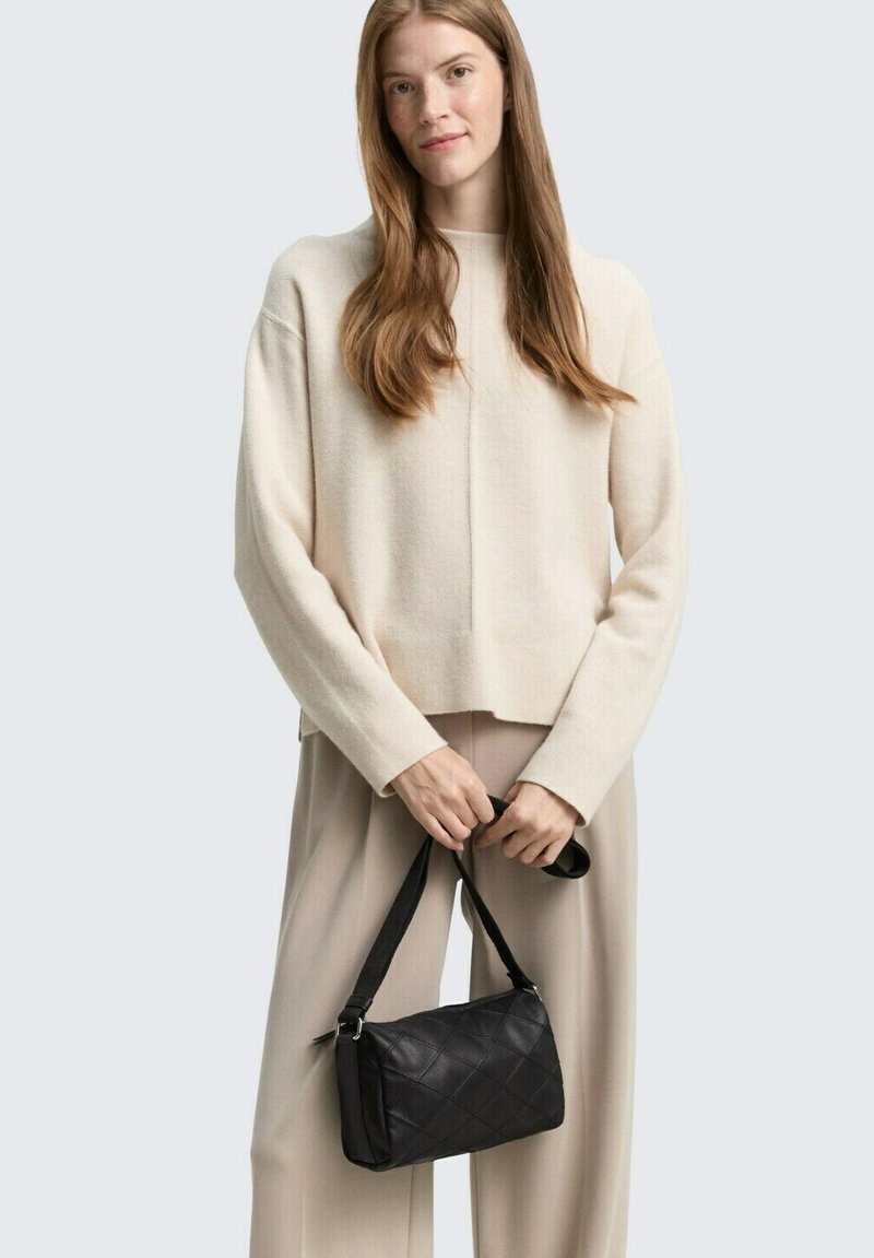 Schwarze gesteppte Handtasche mit einem Tragegurt, in einer Hand gehalten. Cremefarbener Strickpullover und hellbeige Hose, getragen von einem Modell vor einem grauen Hintergrund.