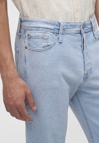 Nahaufnahme einer Person, die hellblaue Jeans und ein strukturiertes weißes Hemd trägt, Hand entspannt an der Seite.