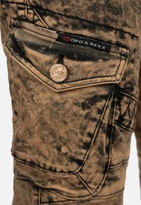 Jeans in denim invecchiato di colore sabbia con motivo a spruzzi neri. Presentano una tasca per le monete con un bottone in metallo marchiato e dettagli cuciti.