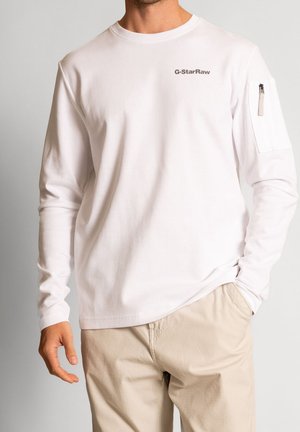 White long-sleeve T-shirt made of textured fabric, featuring a small logo on the chest and a zippered pocket on the left sleeve.