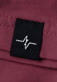 Bordeauxfarbener Stoff mit einem schwarzen Etikett und einem weißen Herzschlag-Design. Die Nähte sind entlang der Kanten sichtbar und betonen Textur und Qualität.