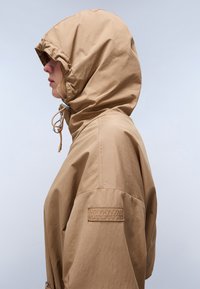 Veste à capuche beige avec une texture légère et lisse ; dotée d'un cordon de serrage réglable au niveau du cou et d'un patch de marque sur la manche.