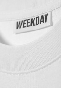 Close-up of a white garment neckline with a white label displaying the word "WEEKDAY" in bold black letters.