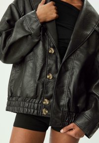 Giacca in pelle sintetica nera con vestibilità ampia, ampi rever e chiusura frontale con bottoni. Presenta orlo a coste e texture con bottoni bronzo.