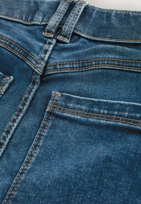 Közepes derékszúrású farmernadrág, kék árnyalatban és sárga varrással; két hátsó zsebbel és sima textúrával rendelkezik.