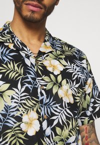 Korte mouwen zwarte shirt met een bloemenpatroon met gele en witte bloemen, groene en blauwe bladeren en bruine knopen.