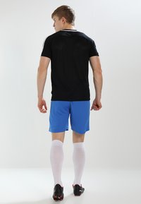 Zwart T-shirt met korte mouwen en witte accenten, gecombineerd met blauwe shorts en witte sokken. Zwarte voetbalschoenen maken de sportieve outfit compleet.