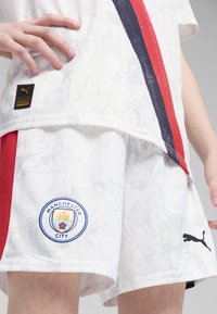 Camiseta y pantalones cortos de fútbol blancos con textura de mármol, rayas rojas y marino, logo del Manchester City y logo negro de Puma.