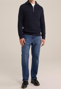 Muž v námořnickém pleteném svetru se zipovým límečkem, v kombinaci s modrými džíny a černými botami, stojí na neutrálním pozadí.