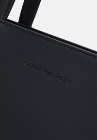 Melnā mākslīgās ādas soma ar reljefu "Calvin Klein Jeans" logotipu. Gluda tekstūra ar kārtīgu šuvēm un minimālistisku dizainu.