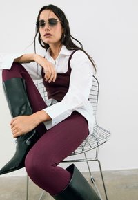 Pantaloni attillati bordeaux, stivali neri in pelle al ginocchio, camicia bianca con bottoni sovrapposta a una canotta scura e occhiali da sole grigi.