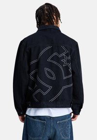 Άτομο με πλεξούδες φοράει μαύρο τζιν μπουφάν και μπλε τζιν παντελόνι, και τα δύο με μεγάλα σχεδιασμένα αστέρια και στυλιζαρισμένα λογότυπα στην πίσω πλευρά.