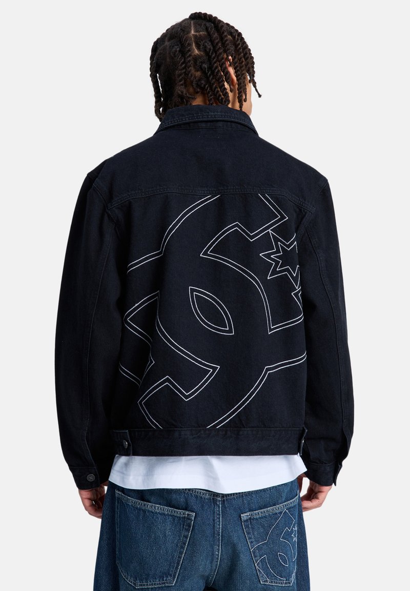 Άτομο με πλεξούδες φοράει μαύρο τζιν μπουφάν και μπλε τζιν παντελόνι, και τα δύο με μεγάλα σχεδιασμένα αστέρια και στυλιζαρισμένα λογότυπα στην πίσω πλευρά.