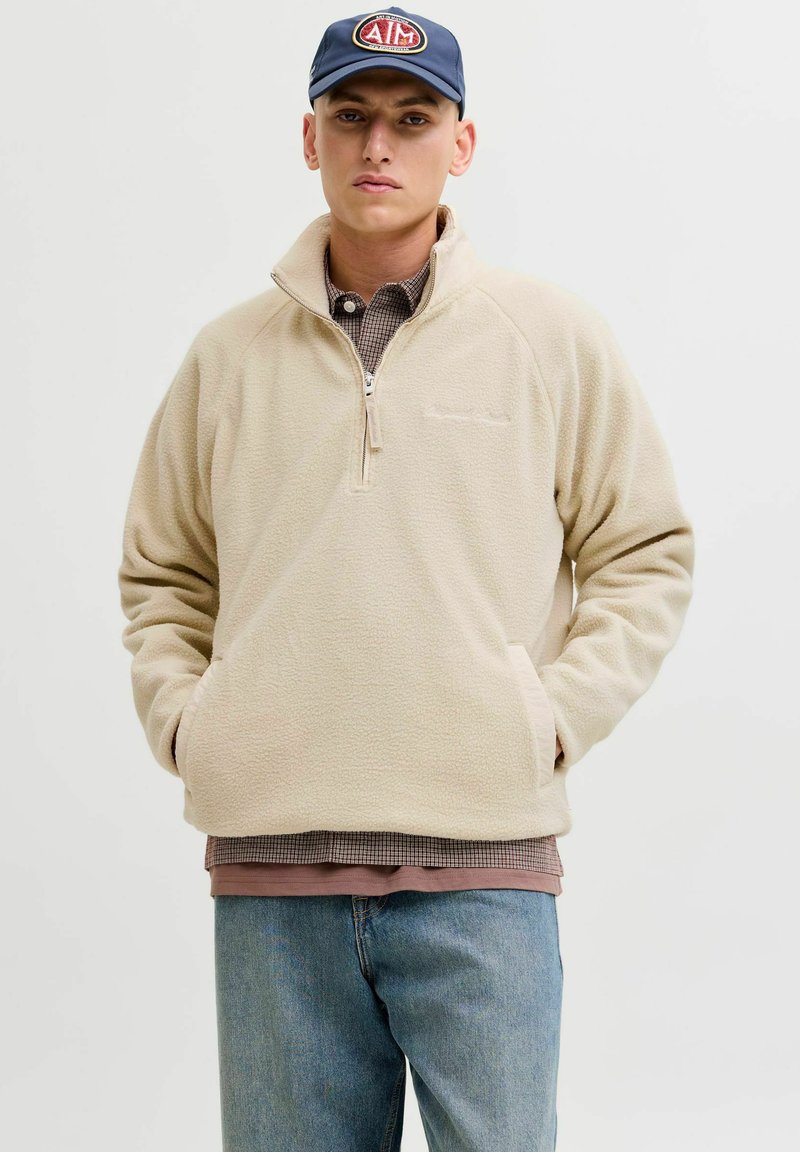 Beige Fleece-Pulloverjacke mit halbem Reißverschluss am Kragen, strukturierter Stoff, Fronttaschen und über einem karierten Hemd getragen. Lässige Jeanshose.