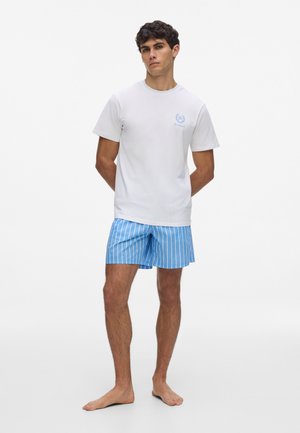 Jeune homme debout pieds nus portant un t-shirt blanc à manches courtes avec un petit logo bleu et un short rayé bleu et blanc.