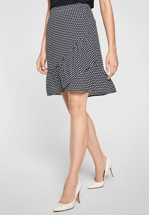 Kvinde iført en sort og hvid nederdel med polkaprikker og flæsekant samt hvide højhælede sko, der går mod en ensfarvet baggrund.