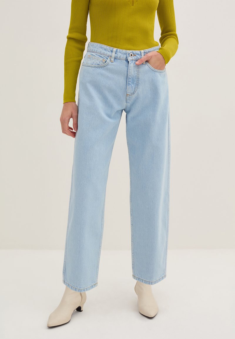 Jeans a gamba larga azzurri chiari con vita alta, dotati di tasche frontali e chiusura con bottone. Abbinati a un top verde oliva a maniche lunghe.