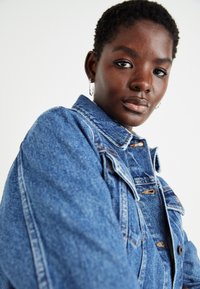 Veste en jean de couleur bleu clair avec une coupe classique, des coutures orange et des boutons en métal. Elle possède un col et deux poches poitrine.