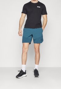 Svart t-shirt med en logotyp, blå shorts med en texturerad panel och fickor, samt svarta träningsskor med vita sulor. Vita strumpor syns.