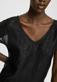 Černý průhledný top s výstřihem do V, s vertikálními metalickými nitěmi. Modelka má na sobě stříbrný řetízek, který zvýrazňuje hladký výstřih.