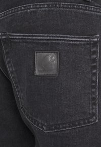 Jeans de mezclilla negra con un bolsillo trasero visible y una etiqueta de marca cuadrada de cuero negro que presenta un logo curvado. La textura es suave con un ligero brillo.