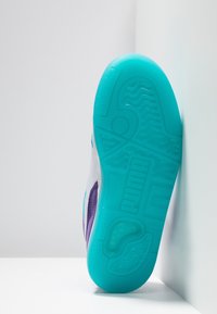 Sole of a sneaker with turquoise tread, white and purple upper, angled upright against a white corner background.