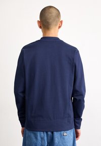 Person med rakad skalle som vänder bort, iklädd en marinblå långärmad tröja och ljusblå denimjeans med en liten lapp på bakfickan.