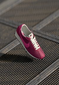 Růžová sportovní bota Nike s texturovaným svrškem, bílými tkaničkami a černou podrážkou, zavěšená nad vzorovaným kovovým povrchem.