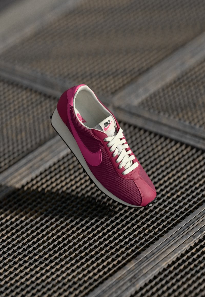 Růžová sportovní bota Nike s texturovaným svrškem, bílými tkaničkami a černou podrážkou, zavěšená nad vzorovaným kovovým povrchem.