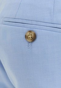 Gros plan sur un tissu bleu clair présentant un seul bouton beige à quatre trous, un détail de couture et le bord d'une poche soigneusement cousu.