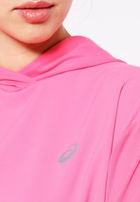 Růžová mikina s kapucí, vyrobená z lehkého a hladkého materiálu, s stříbrným logem na hrudi a s tkaničkou na stažení kapuce.