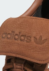 Vrchná časť tenisky z hnedej kože s textúrovaným povrchom, zdobená embosovaným logom "adidas" a logom so tromi pruhmi. Textúrovaný látkový golier a šnúrky.