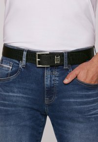 Schwarzer Gürtel aus Krokodilleder mit silberner Metall-Schließe, getragen über blauen Jeans mit heller Auswaschung und kontrastierenden Nähten.