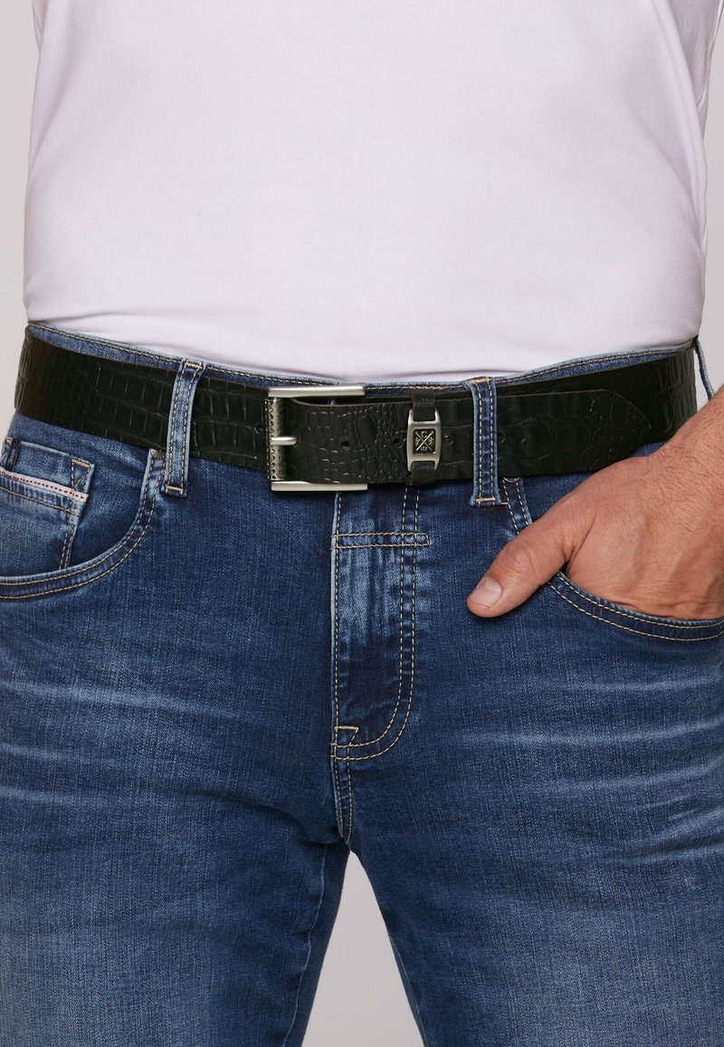 Schwarzer Gürtel aus Krokodilleder mit silberner Metall-Schließe, getragen über blauen Jeans mit heller Auswaschung und kontrastierenden Nähten.