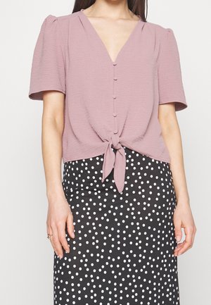 Blusa mauve a maniche corte con scollo a V, dettagli in bottoni e davanti annodato, abbinata a una gonna nera con pois bianchi.