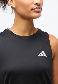 Κοντινό πλάνο ατόμου που φοράει μαύρο αθλητικό τοπ χωρίς μανίκια με λευκό λογότυπο Adidas στο στήθος, με φόντο άσπρο.