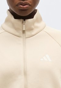 Εγγύς εικόνα ατόμου που φοράει μπεζ ζακέτα με φερμουάρ και ψηλό γιακά, με το λογότυπο της Adidas στο στήθος.