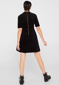 Schwarzes Cordkleid mit kurzen Ärmeln, einem taillierten Oberteil und einem ausgestellten Rock, ausgestattet mit einem zentralen Reißverschluss auf der Rückseite mit einem metallicfarbigen Ziehgriff.