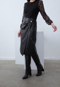 Schwarzer Lederrock mit asymmetrischem Saum, der einen seitlichen Schlitz und ein Knopfdetail aufweist, kombiniert mit einem schwarzen Oberteil mit transparenten Punktenärmeln.