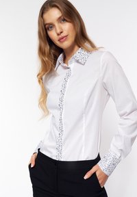 Biała koszula zapinana na guziki z kwiatowym wzorem przy kołnierzyku i mankietach. Tkanina jest gładka, a krój dopasowany z długimi rękawami.