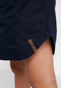 Jupe bleu marine avec ourlet incurvé et étiquette en tissu marron, portée au-dessus du genou montrant une partie d'une jambe nue.