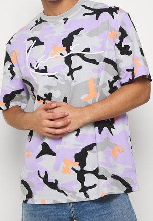 Personne portant un t-shirt camouflage à manches courtes avec des formes violettes, noires, orange et grises et un texte blanc en écriture cursive sur la poitrine.