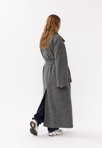 Cappotto lungo grigio di lana con vita cinturata, maniche ampie e spacco posteriore. Il modello indossa jeans e sneakers. Lo sfondo è bianco uniforme.