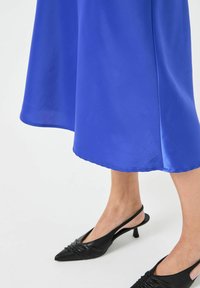 Satino mėlynas suknelė su laisvu siluetu, turinti hemą, kuris nukrenta iki vidurio blauzdos. Suderinta su juodomis, smailianosiomis slingback batų su raukšlėmis.