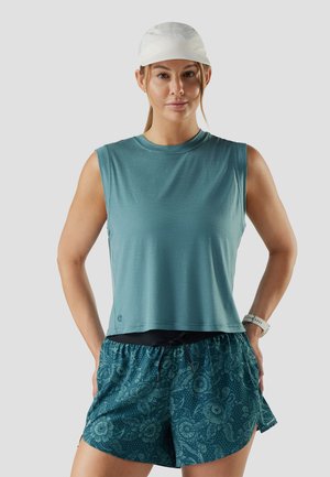 Donna con top senza maniche turchese, pantaloncini turchese a motivo floreale, cappellino sportivo bianco e smartwatch bianco, in piedi con le mani sui fianchi su sfondo grigio.