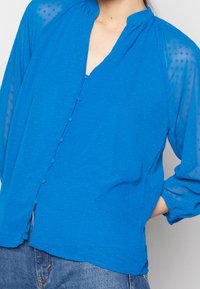 Blå blus med knappstängning fram, texturerade prickar, lättviktsmaterial och upprätt krage. Löst sittande, långa genomskinliga ärmar.